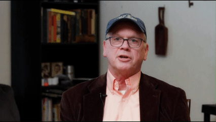 Man with a hat and peach shirt with a bookshelf in the background.